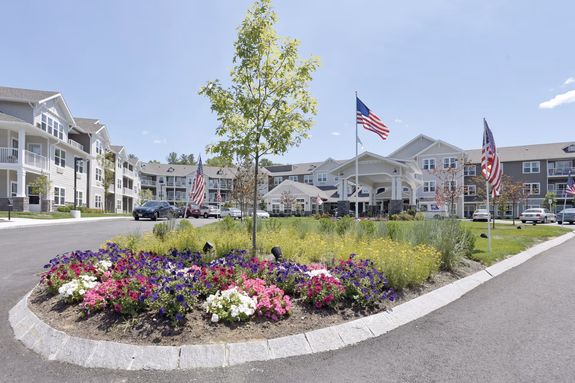 Front entrance of a multi-story senior living building with a landscaped flower roundabout, American flags, and parked cars.