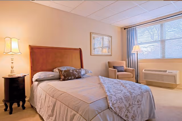 A cozy bedroom with a wooden headboard bed made with beige bedding and decorative pillows. There is a nightstand with a lamp on the left side of the bed. A beige armchair with a dark cushion is placed near a window with blue curtains and blinds. A floor lamp stands next to the armchair, and a wall-mounted air conditioning unit is below the window. A framed picture hangs on the wall above the armchair.