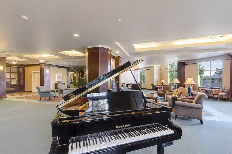 A spacious and well-lit common area in a senior living facility featuring a black grand piano in the foreground. The room has comfortable seating arrangements including armchairs and sofas, wooden columns, large windows allowing natural light, and table lamps providing additional lighting.