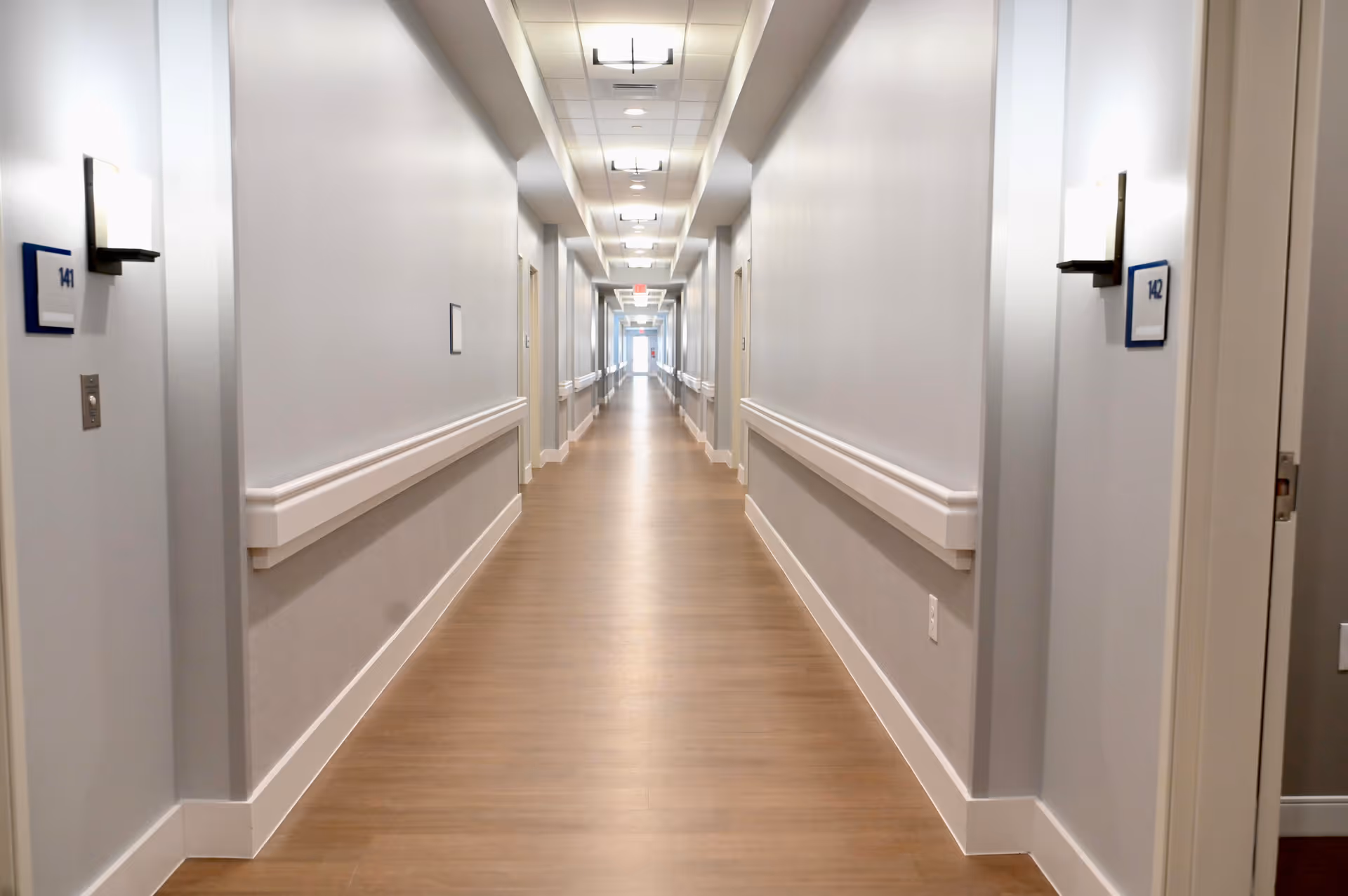 A long, well-lit interior corridor with handrails, room number plaques, and wood-look flooring in a senior living facility.