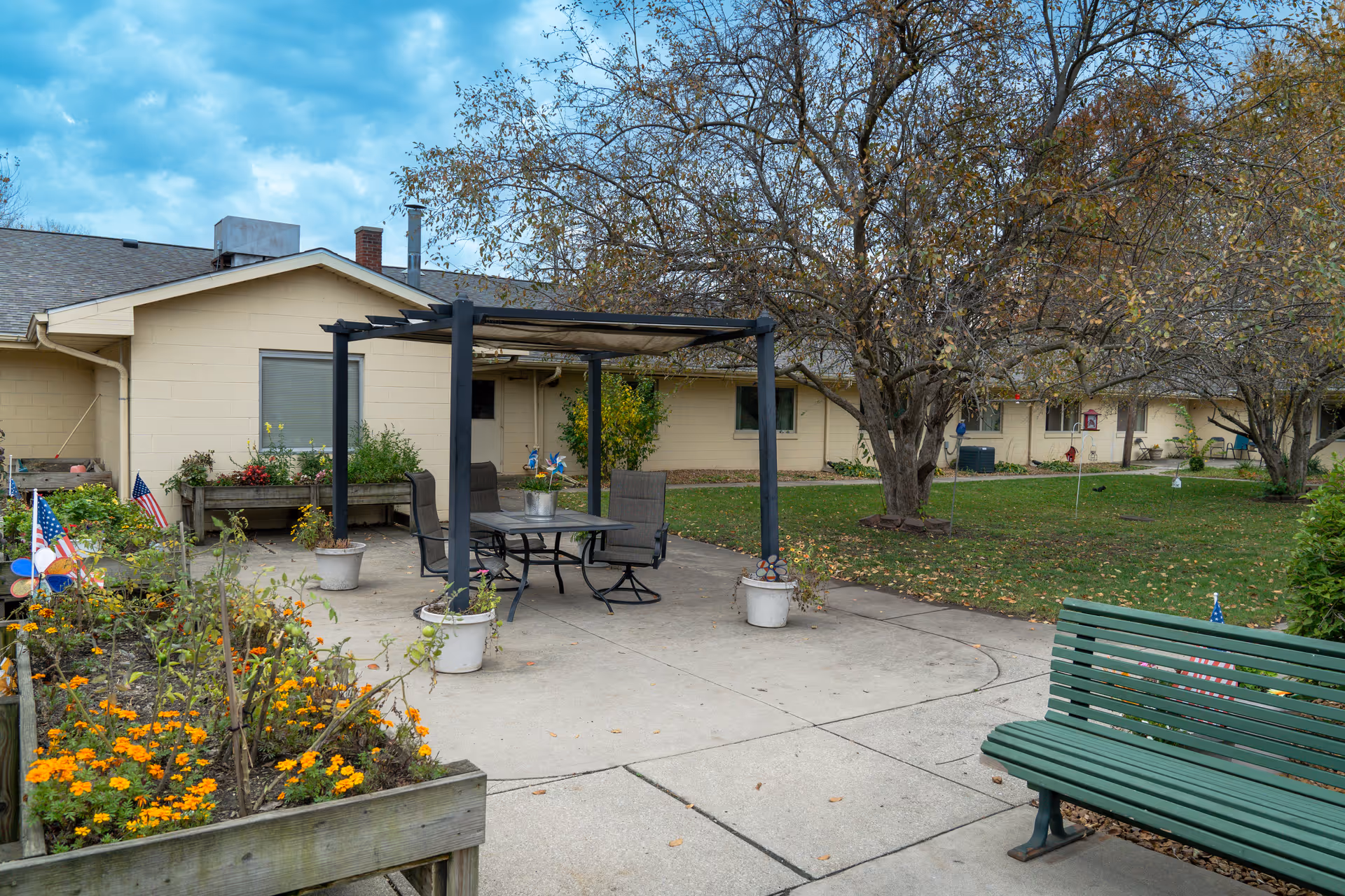 Outdoor patio area at Warsaw Meadows Care Center featuring a pergola with a table and four chairs, surrounded by potted plants and flower beds. There is a green bench on the right side and trees with sparse leaves in the background. The building is a single-story structure with beige walls and several windows.