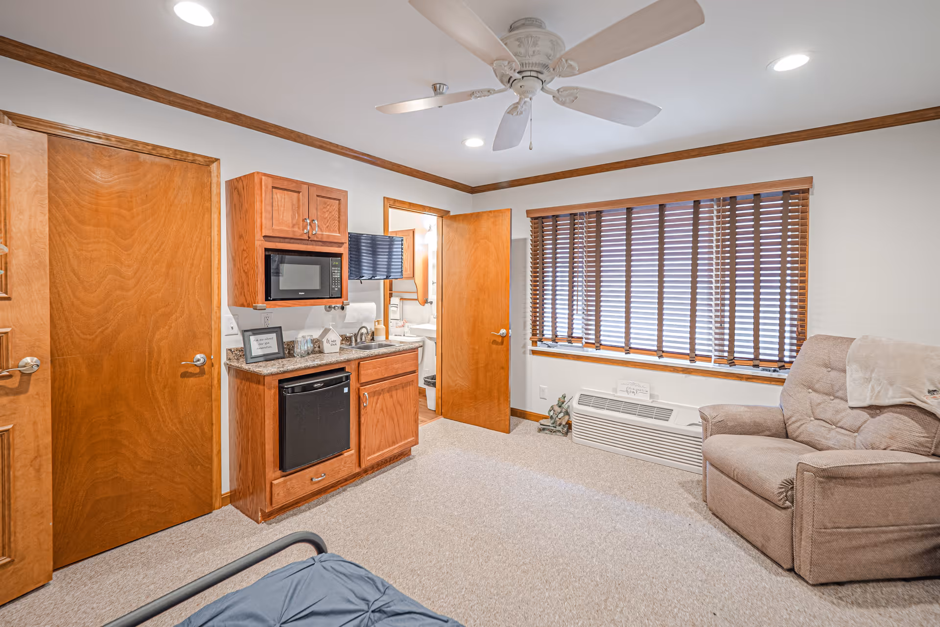 A cozy room in a senior living facility featuring a beige recliner chair with a white throw on the right, a window with wooden blinds, a ceiling fan with lights, and a kitchenette area with a microwave, mini fridge, and sink. There are two wooden doors, one slightly open revealing a bathroom with a sink and toilet.