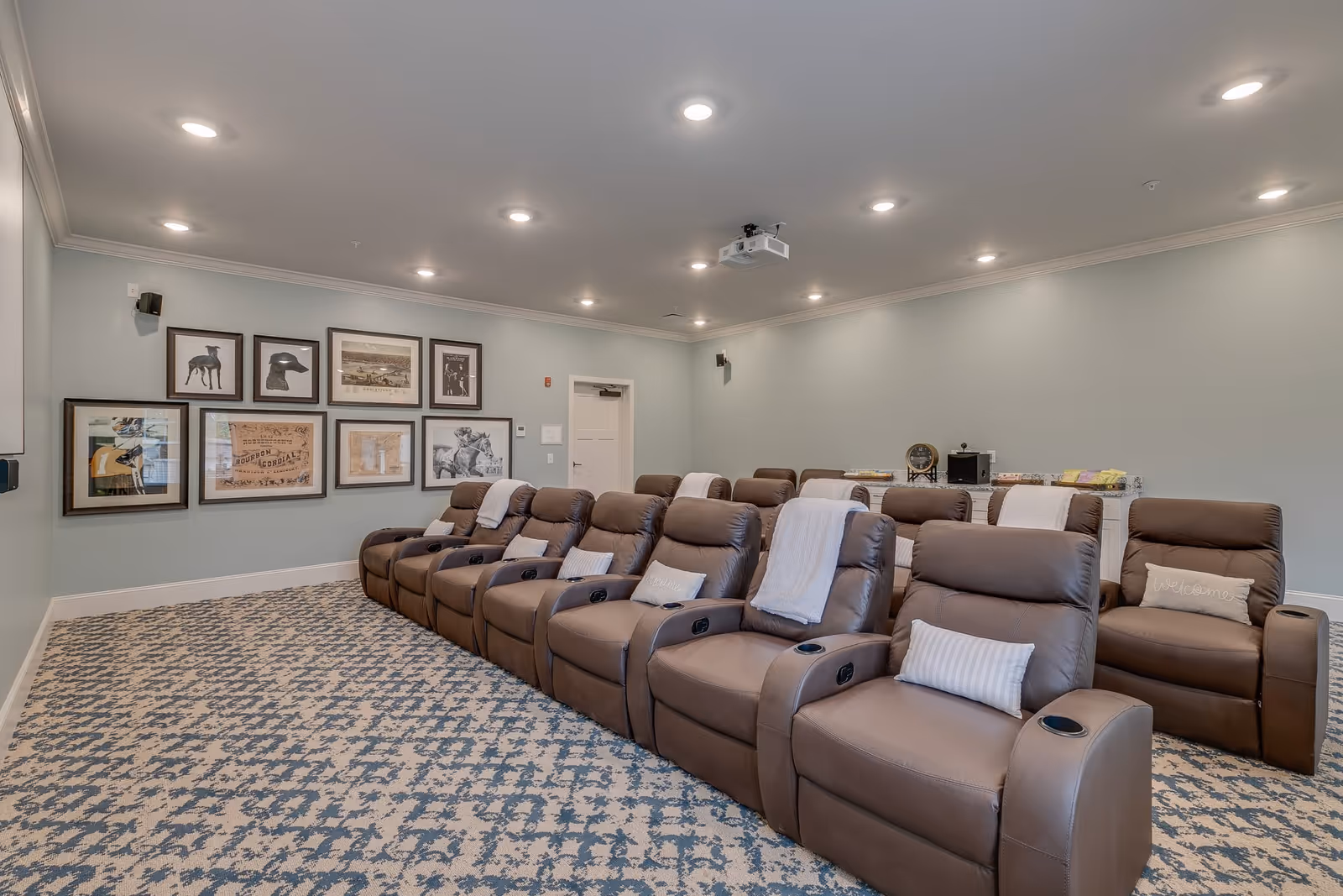 A cozy theater room with two rows of brown leather recliner chairs, each equipped with cup holders. The chairs have white and striped pillows and some have white throws. The room has light blue walls adorned with framed artwork and a patterned carpet. Ceiling lights and a projector are visible.