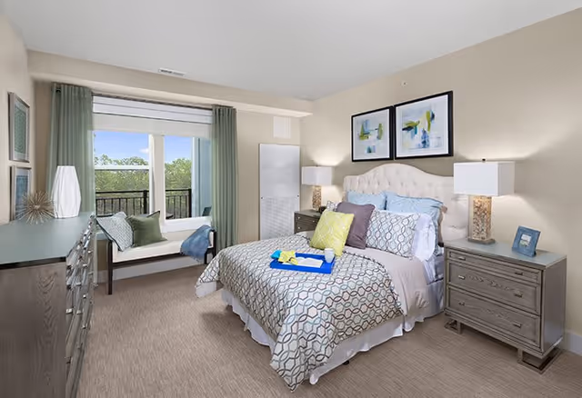 A bright and cozy bedroom with a large bed featuring patterned bedding and multiple pillows. There are two nightstands with lamps on either side of the bed. Above the bed, two framed abstract paintings hang on the wall. A window seat with cushions is positioned next to a large window with green curtains, allowing natural light to fill the room. A dresser with decorative items is on the left side of the room.