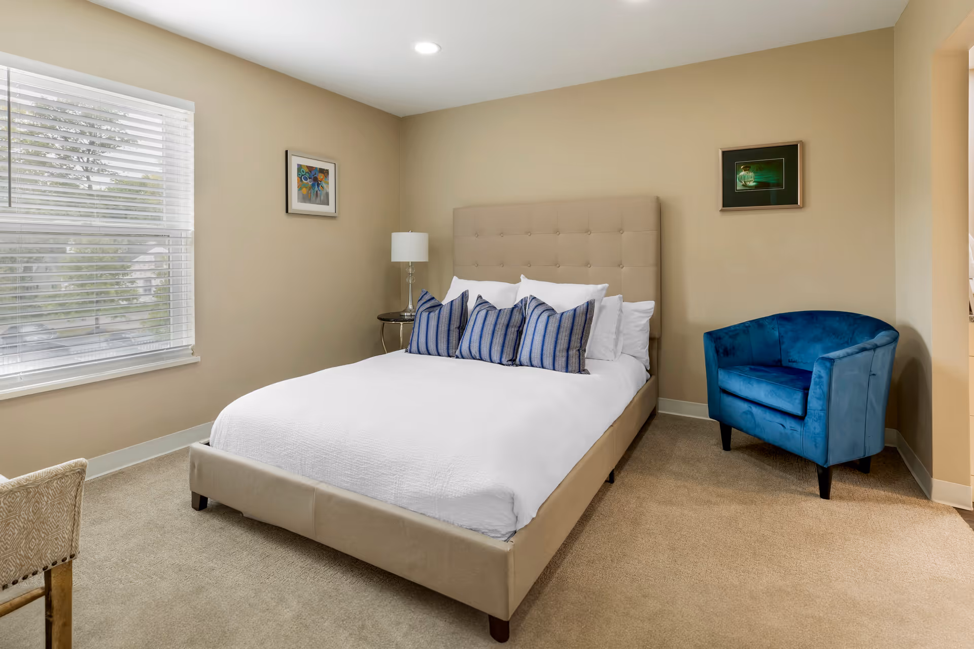 A bedroom with a beige upholstered bed featuring white bedding and four blue patterned pillows. There is a blue armchair in the corner, a small round side table with a lamp, beige walls with two framed pictures, and a window with white blinds letting in natural light.