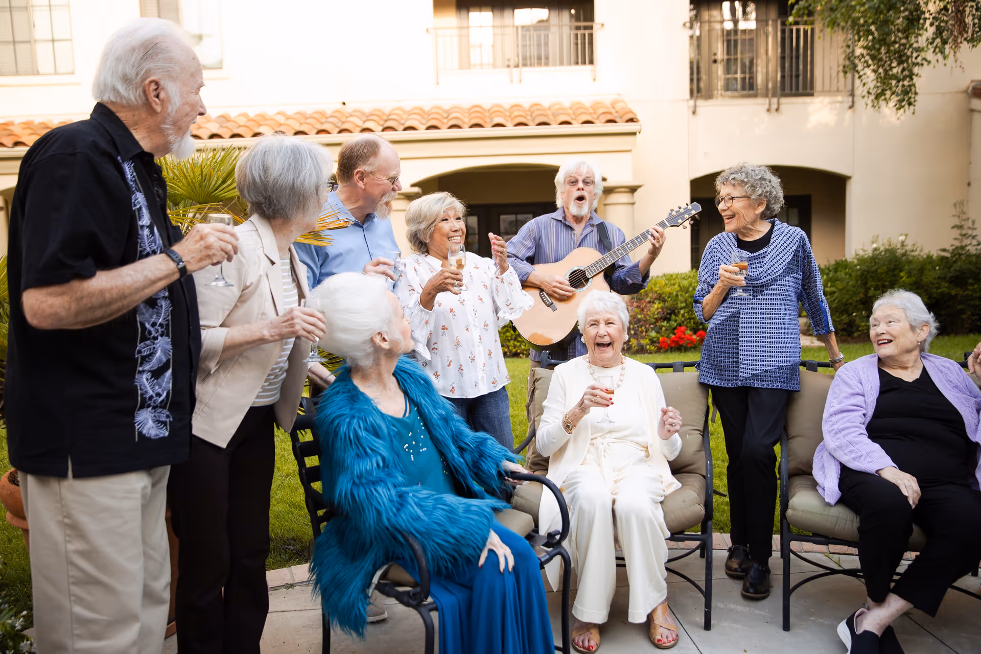 A group of elderly people gathered outdoors in a garden area of a senior living facility. They are smiling, laughing, and holding drinks while a man plays an acoustic guitar. The setting is cheerful and social, with greenery and a building with balconies in the background.