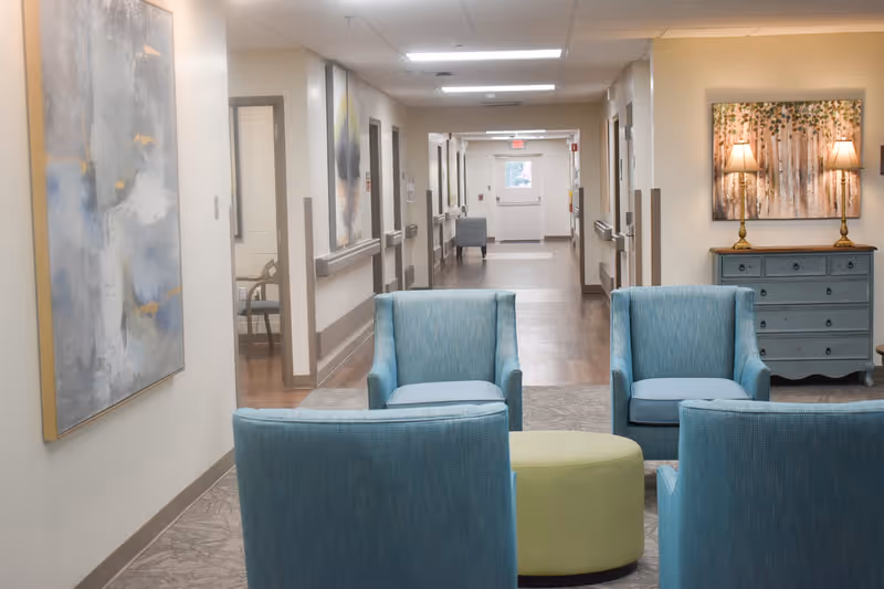 A hallway seating area in a senior care facility with four teal armchairs around a green ottoman, artwork on the walls and a dresser with lamps.