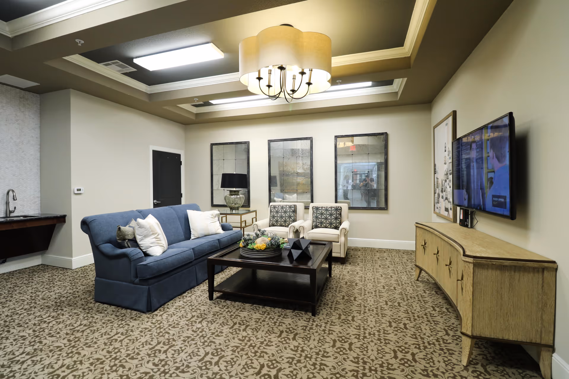 A cozy living room area in an assisted living facility featuring a blue sofa with white and patterned pillows, two beige armchairs with patterned cushions, a dark wooden coffee table with decorative items, a side table with a lamp, three large framed mirrors on the wall, a wall-mounted TV above a wooden console, and a patterned carpet. The ceiling has a recessed design with a large light fixture.