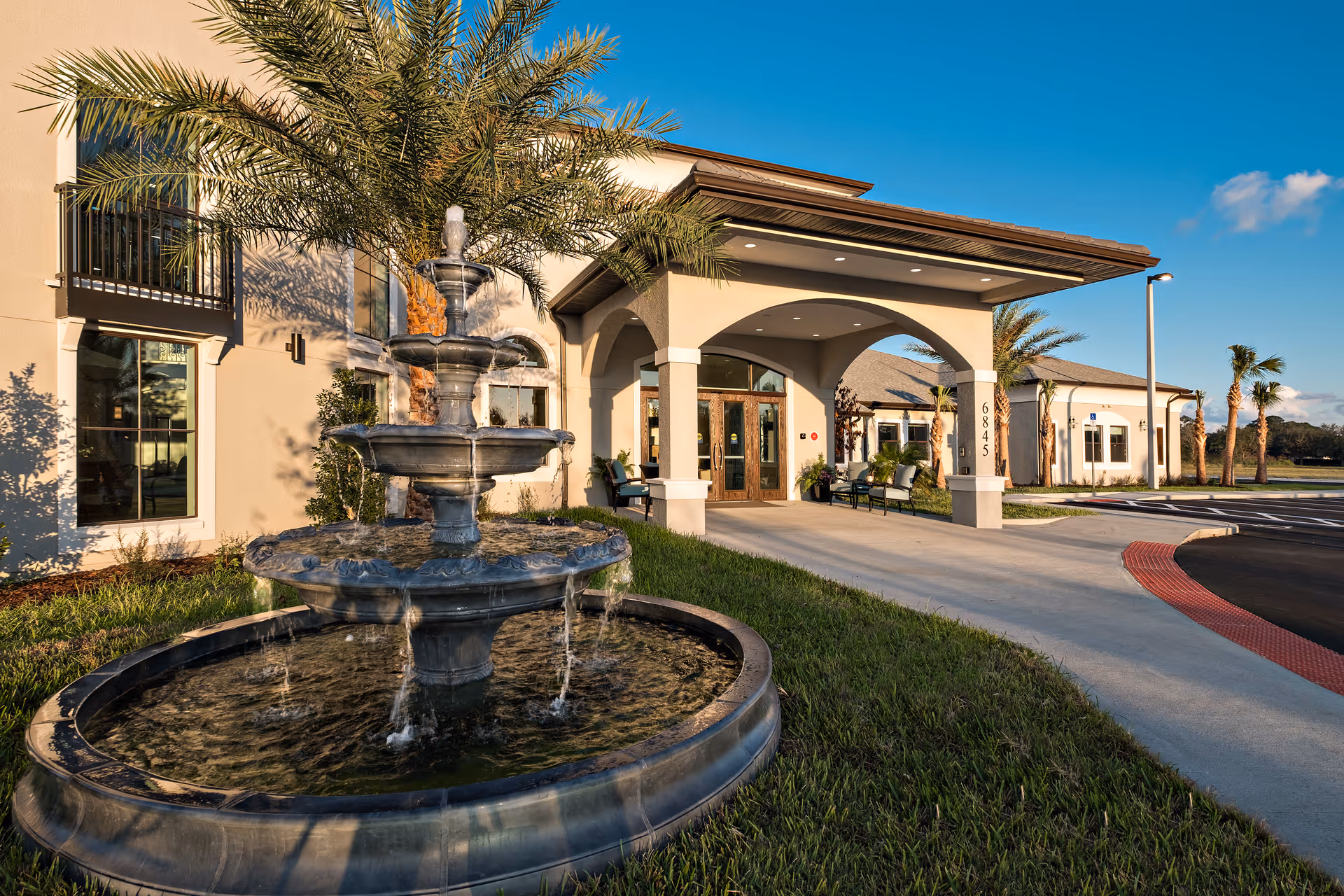 Exterior view of Market Street Viera facility entrance with a multi-tiered water fountain in the foreground, palm trees, benches near the entrance, and a clear blue sky.