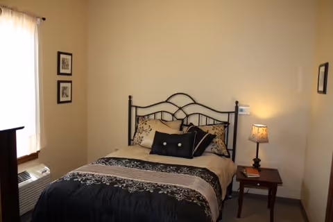 A bedroom with a black metal bed frame, a bed made with black, beige, and patterned bedding, a wooden nightstand with a lamp and a book, two framed pictures on the wall, and a window with white curtains.