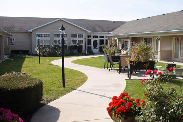 Outdoor courtyard area of Kokomo Place senior living facility with a curved concrete walkway, green grass, flower beds with red flowers, outdoor seating with chairs and tables, and beige buildings surrounding the courtyard under a clear sky.