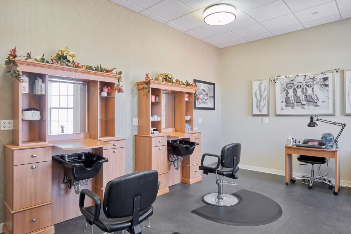 Interior view of a salon area in a senior living facility with two wooden styling stations, each equipped with a black wash basin and a black salon chair. The stations have shelves with various hair care products and decorative greenery on top. There is a small desk with a chair and a lamp against the far wall, which is decorated with framed black and white artwork.