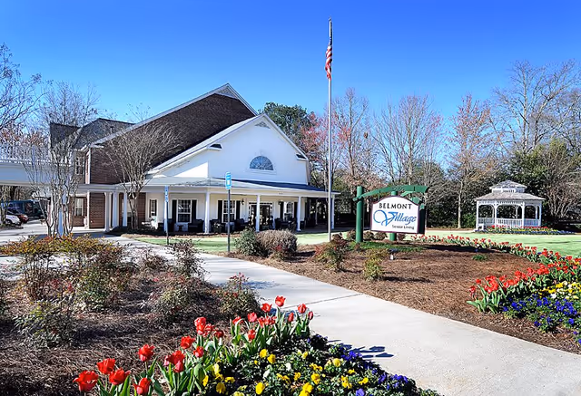 Exterior view of Belmont Village Senior Living Buckhead facility with a clear blue sky, a garden with colorful flowers, a paved walkway, an American flag on a pole, and a white gazebo in the background.