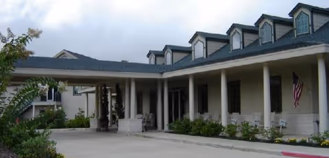 Exterior view of a senior living facility with a covered entrance supported by columns, multiple dormer windows on the roof, and an American flag hanging near the entrance. There are shrubs and greenery along the building and a driveway in front.