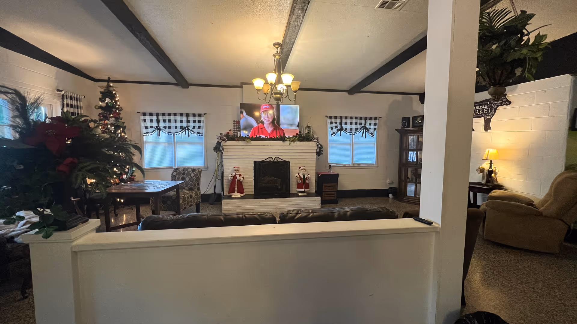 A cozy living room area decorated for Christmas with a Christmas tree on the left, a white brick fireplace in the center with holiday figurines and garland, a TV mounted above the fireplace showing a woman in a red uniform, and comfortable seating including a leather couch and an armchair. The room has white walls with dark wooden beams on the ceiling and checkered curtains on the windows.