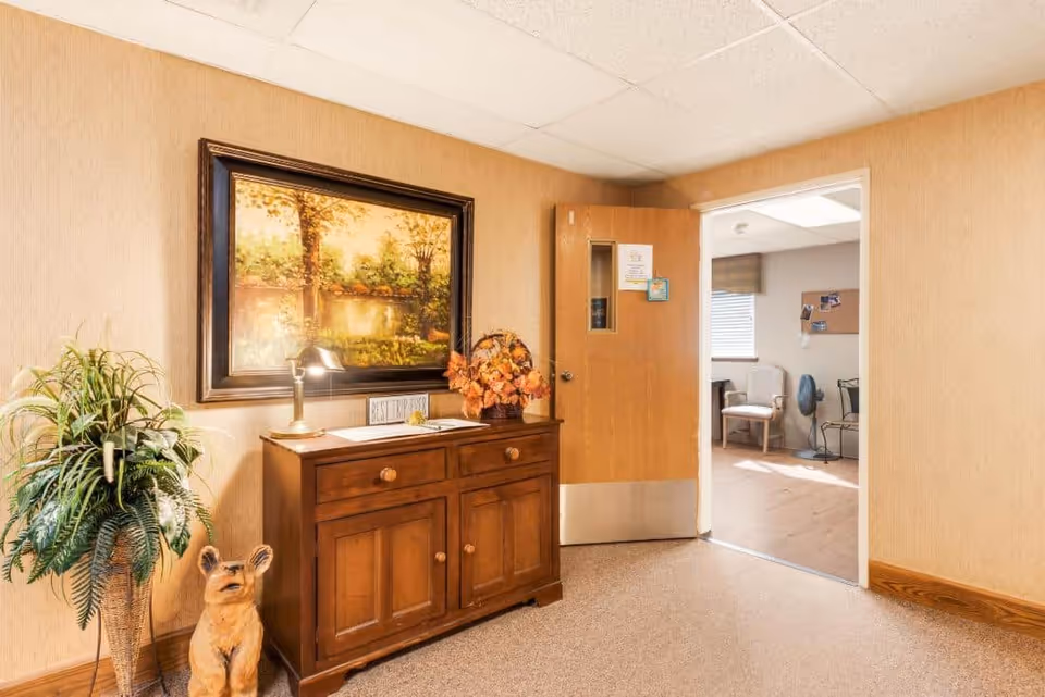 A warmly lit interior hallway with beige walls and carpeted floor. A wooden cabinet with drawers and doors is against the wall, decorated with a lamp, a basket of artificial flowers, and a framed landscape painting above it. To the left of the cabinet is a tall potted plant and a small dog statue. An open wooden door leads to another room with chairs, a fan, and a bulletin board visible.