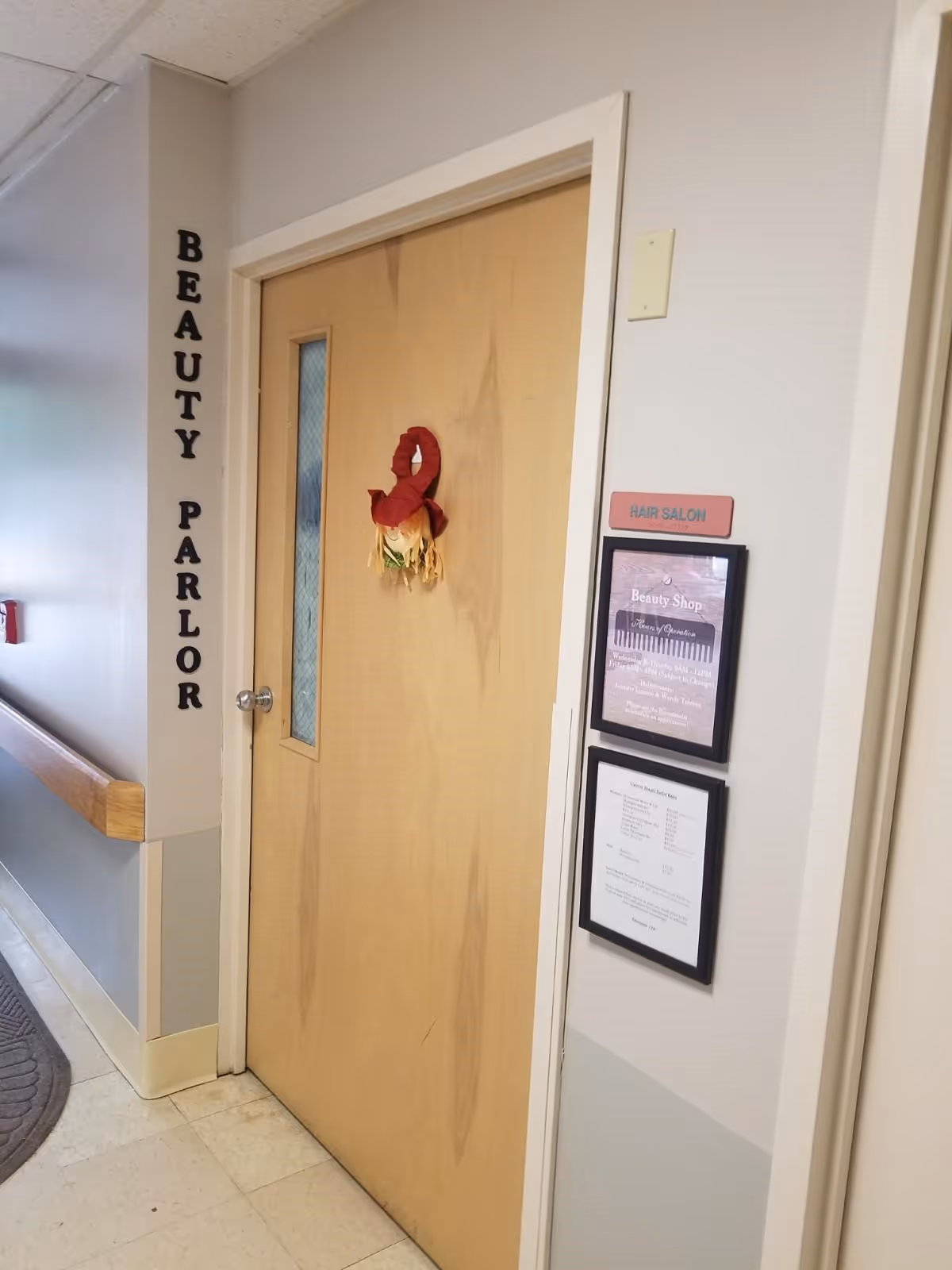 A hallway in a senior living facility with a wooden door labeled 'Beauty Parlor' and a small decorative item hanging on the door. The wall next to the door has a sign that reads 'Hair Salon' and two framed notices below it. The hallway has a handrail and a floor mat visible on the left side.