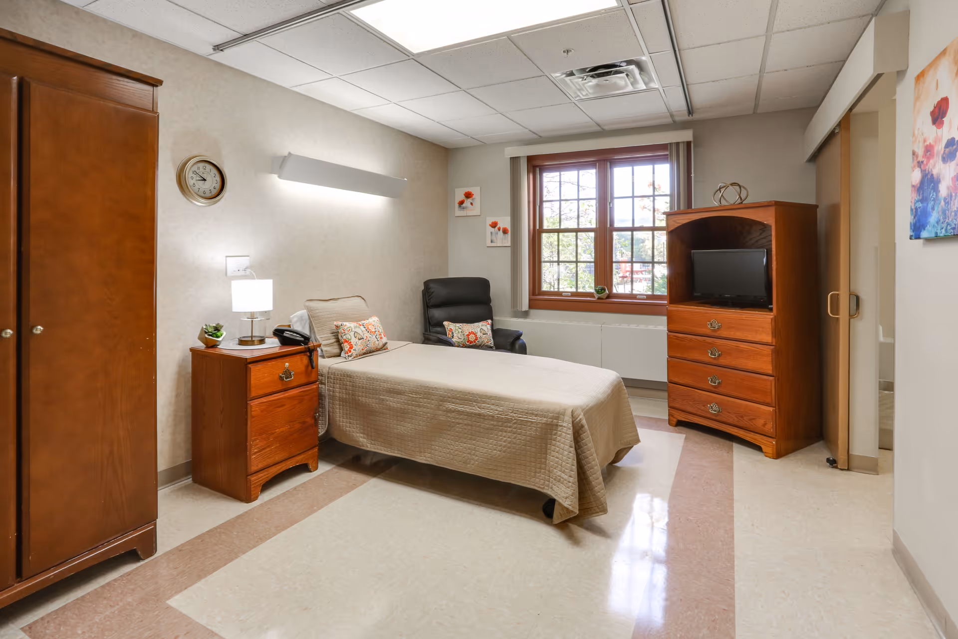 A simple, clean bedroom in a senior living facility featuring a single bed with a beige quilt, a wooden nightstand with a lamp and a phone, a black armchair with a decorative pillow, a wooden wardrobe, a wooden dresser with a small TV, and a window with a view outside. The walls are light-colored with minimal artwork, and the floor has a beige and brown pattern.