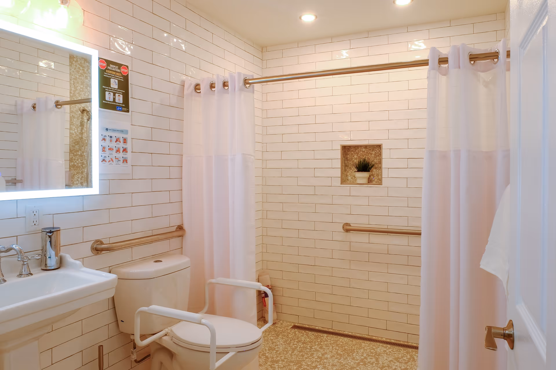 A clean and accessible bathroom with white tiled walls and a beige patterned floor. The bathroom features a white toilet with safety rails, a white pedestal sink with a chrome faucet, a large mirror with built-in lighting, and a shower area with a white curtain and grab bars. There is a small recessed shelf in the shower wall holding a small potted plant.