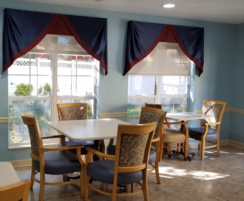 A dining area with two white square tables surrounded by wooden chairs with patterned backs and blue cushions. The room has light blue walls and two large windows with blue and maroon valances, letting in natural sunlight. Outside the windows, greenery and a white fence are visible.