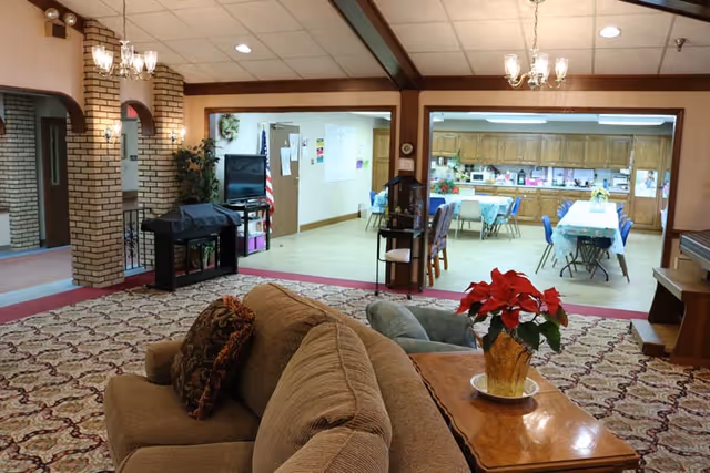 Interior view of a senior living facility showing a cozy living room area with a brown couch and a wooden coffee table with a red poinsettia plant. In the background, there is a dining area with tables and chairs, and a kitchen with wooden cabinets. The room has brick archways, chandeliers, and a patterned carpet.