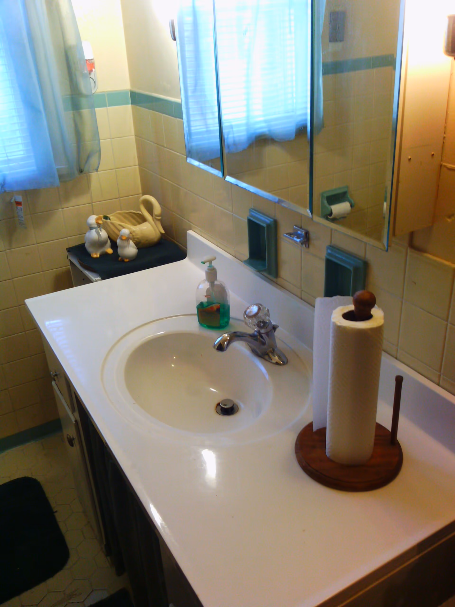 Bathroom sink area with a white countertop, a chrome faucet, a bottle of green liquid soap, and a wooden paper towel holder with a roll of paper towels. The wall behind the sink is tiled with beige tiles and has a large mirror above it. To the left, there is a small table with decorative ceramic ducks and a window with sheer curtains.