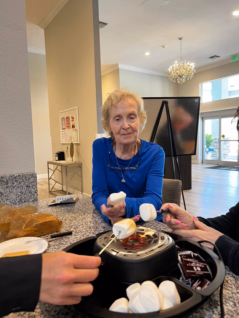 An elderly woman in a blue shirt is roasting marshmallows on a small indoor electric grill at a granite countertop. Two other people are also holding marshmallows over the grill. The setting appears to be a bright, modern common area with a chandelier and large windows in the background.
