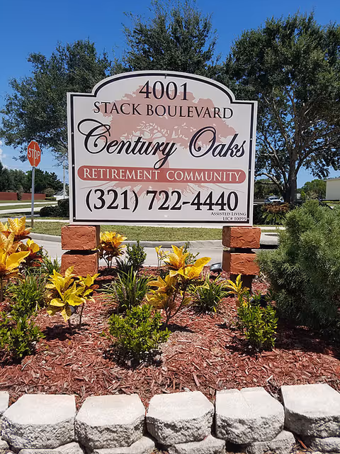 Outdoor sign for Century Oaks Retirement Community at 4001 Stack Boulevard, surrounded by landscaping with small bushes and plants, with trees and a stop sign in the background under a clear blue sky.