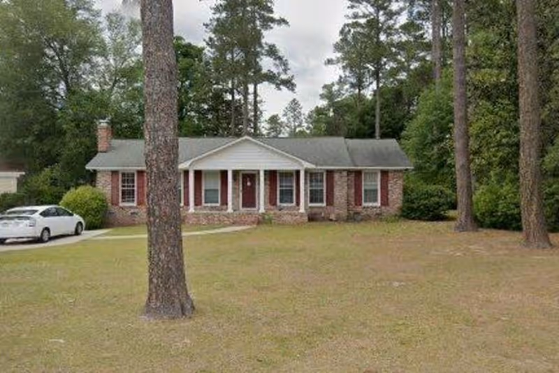 Single-story brick house with a gray roof and red shutters, surrounded by tall pine trees and a large grassy yard. A white car is parked on the driveway to the left of the house.