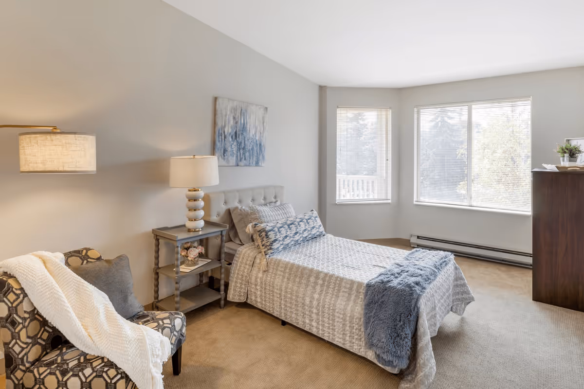 A bright and cozy bedroom with a single bed covered in a light gray and blue patterned bedspread, a blue throw blanket, and matching pillows. Next to the bed is a small gray nightstand with a lamp and decorative items. A patterned armchair with a white throw blanket is positioned near the nightstand. The room has beige carpeting, light gray walls, and two large windows letting in natural light. A dark wooden dresser with a plant and books is placed against the wall near the windows.