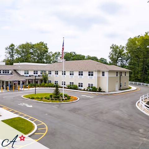 Exterior view of a two-story assisted living facility building with beige siding and a gray roof. The building is surrounded by a paved driveway with a circular landscaped island featuring a flagpole with an American flag. Trees and greenery are visible in the background under a partly cloudy sky.