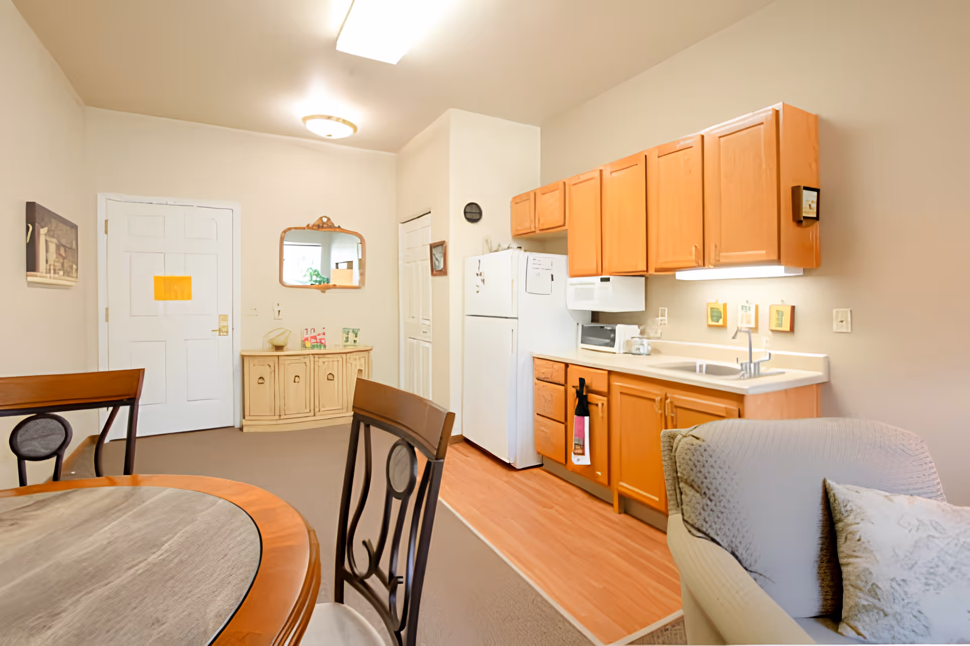 Interior view of a senior living facility kitchen and dining area featuring wooden cabinets, a white refrigerator, microwave, and sink. A round dining table with chairs is partially visible in the foreground, and a cushioned armchair with a pillow is on the right. The walls are light-colored with a mirror and a small cabinet near the door.