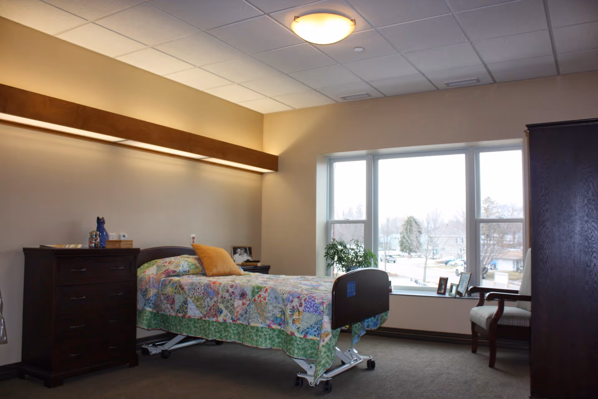 A well-lit bedroom in a senior living facility with a single hospital-style bed covered with a colorful patchwork quilt and a yellow pillow. The room has a large window with a view of trees and houses outside, a wooden dresser with decorative items, a wooden wardrobe, and a cushioned armchair near the window.