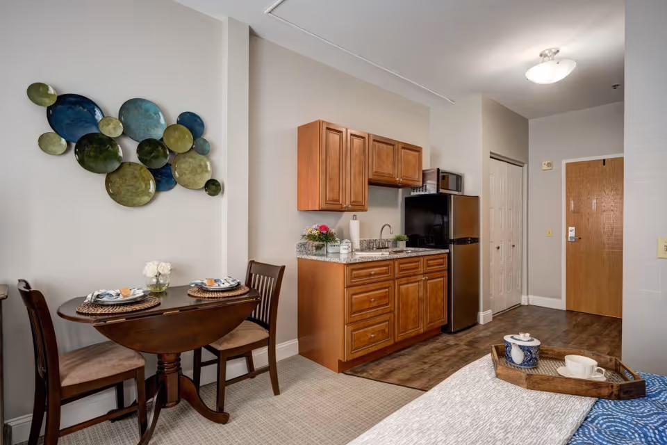 A cozy studio apartment interior featuring a small wooden dining table set for two with plates and glasses, a kitchenette with wooden cabinets, a granite countertop, a sink, a microwave, and a refrigerator. The room has light-colored walls with decorative circular wall art, wood flooring near the kitchenette, and carpeted flooring near the dining area. A wooden door and a closet with white double doors are visible in the background. A tray with a teapot and cup is placed on a bed in the foreground.