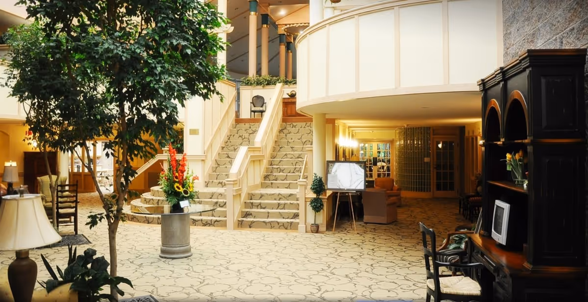 Spacious and well-lit interior common area of a senior living facility with a large staircase in the center, decorative plants, floral arrangements, comfortable seating, and wooden furniture including a desk with a computer monitor.