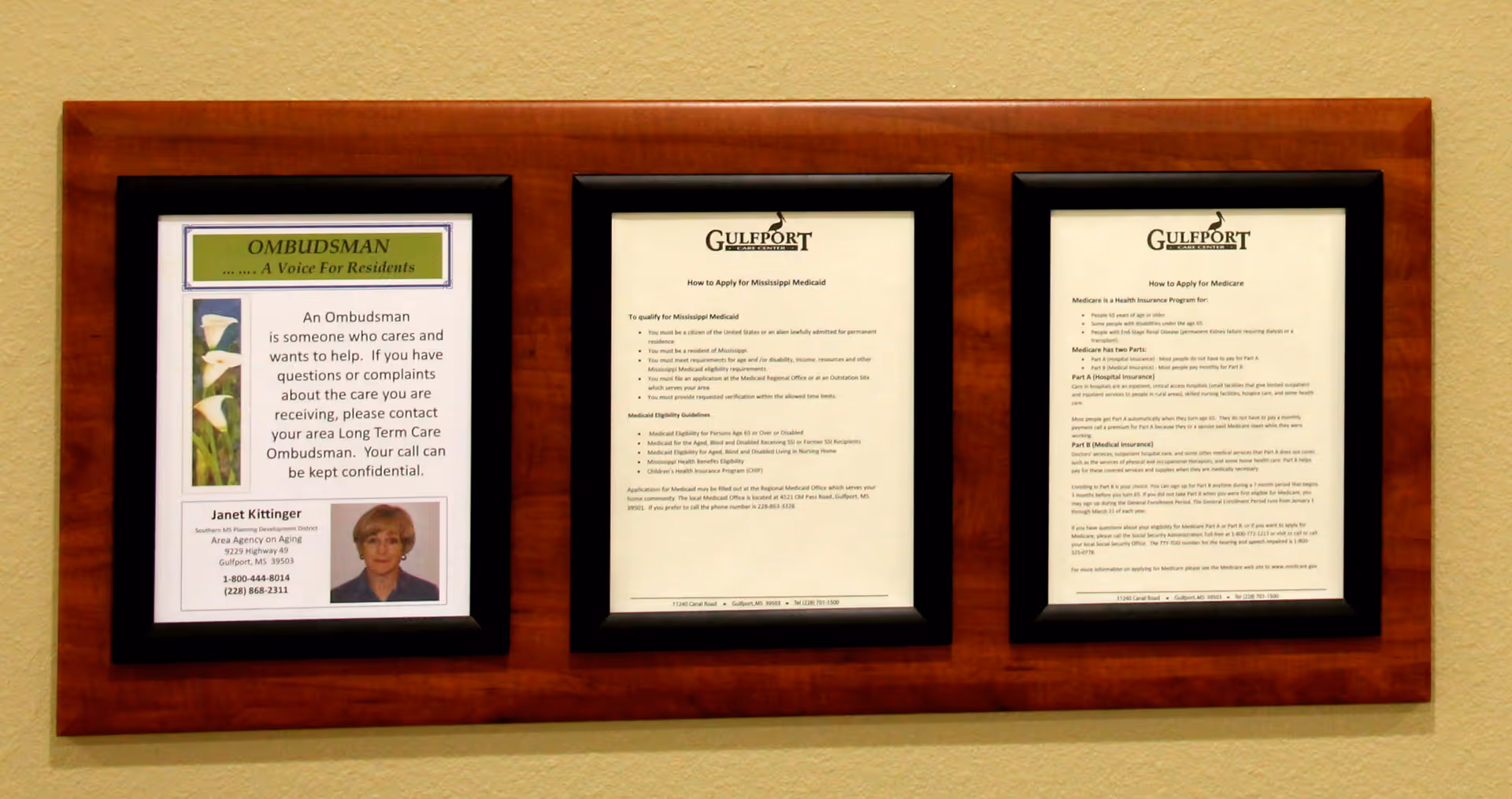Three framed informational notices mounted on a wooden plaque on a beige wall. The left frame provides contact information for an Ombudsman named Janet Kittinger, the middle frame explains how to apply for Mississippi Medicaid, and the right frame details how to apply for Medicare.