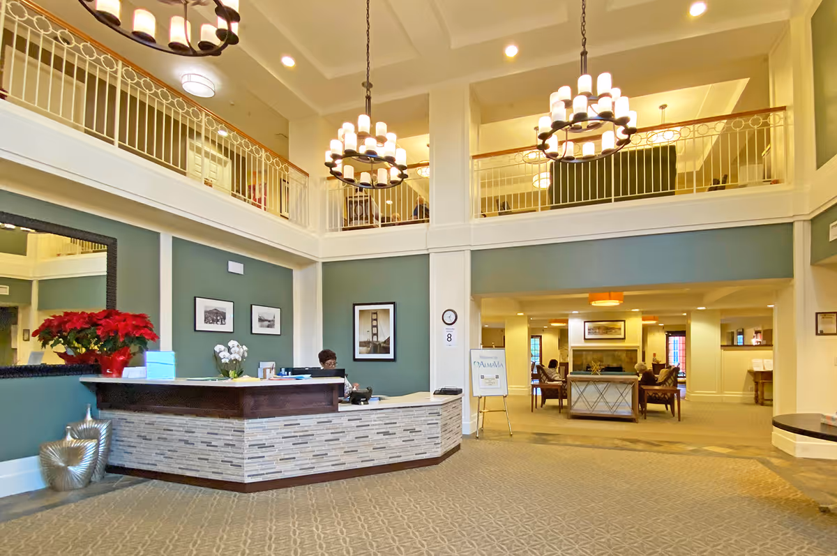 Spacious senior living facility lobby with a reception desk on the left, decorated with poinsettia plants and flowers. The area has high ceilings with two large chandeliers, a balcony railing on the upper floor, and a seating area with people sitting near a fireplace in the background.