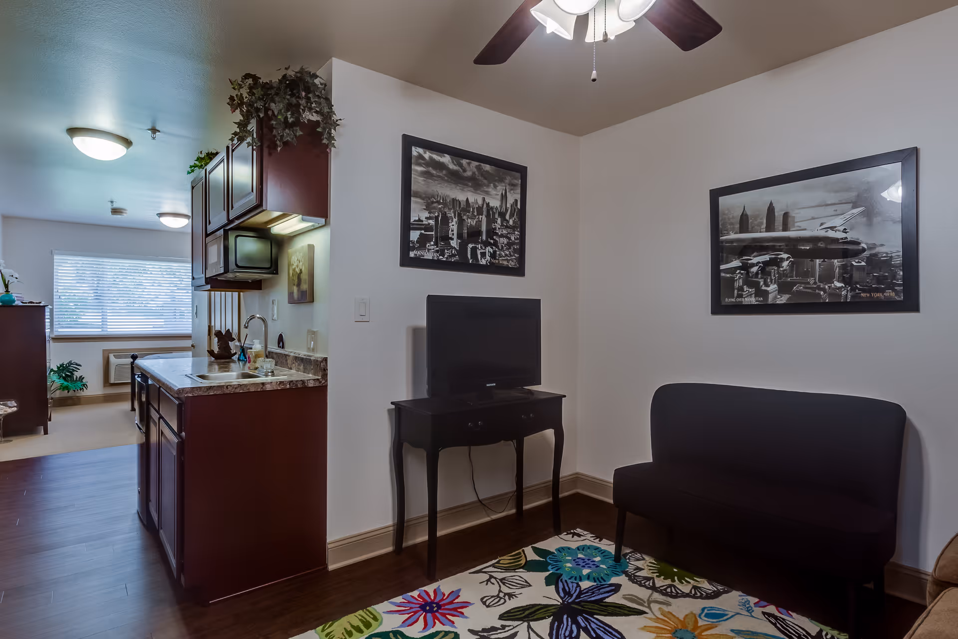 Interior view of a senior living facility apartment showing a small living area with a black loveseat, a TV on a small black table, and two black and white framed pictures on the wall. To the left is a kitchenette with dark wood cabinets, a countertop with a sink, and a microwave. In the background, there is a window with blinds and some plants near a heating unit.