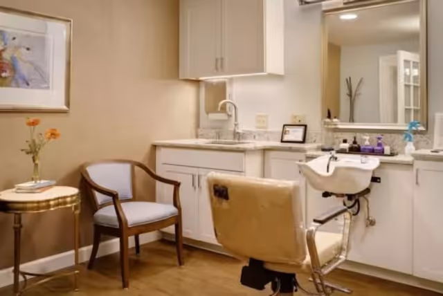 A small salon area in a senior living facility featuring a salon chair in front of a sink and mirror, a wooden armchair with light upholstery, a small round side table with a vase of flowers and a book, and white cabinetry with a countertop.