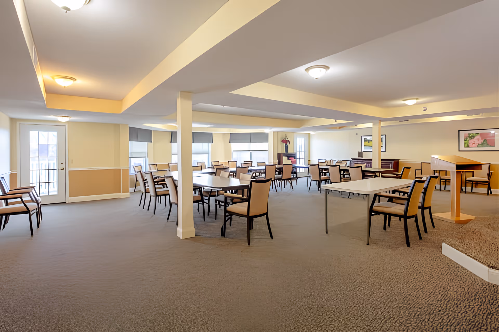 Spacious communal dining/activity room with multiple tables, chairs, and a small podium in a senior living facility.