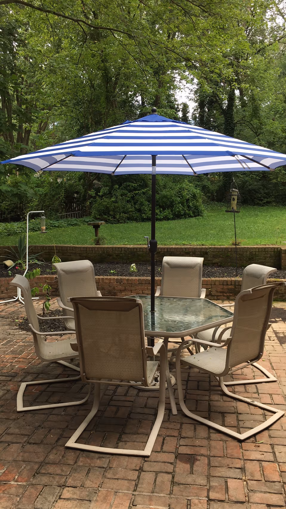 Outdoor patio area with a glass-top table surrounded by six beige mesh chairs. A large blue and white striped umbrella is centered over the table. The patio is made of brick and is adjacent to a grassy area with trees and bird feeders in the background.