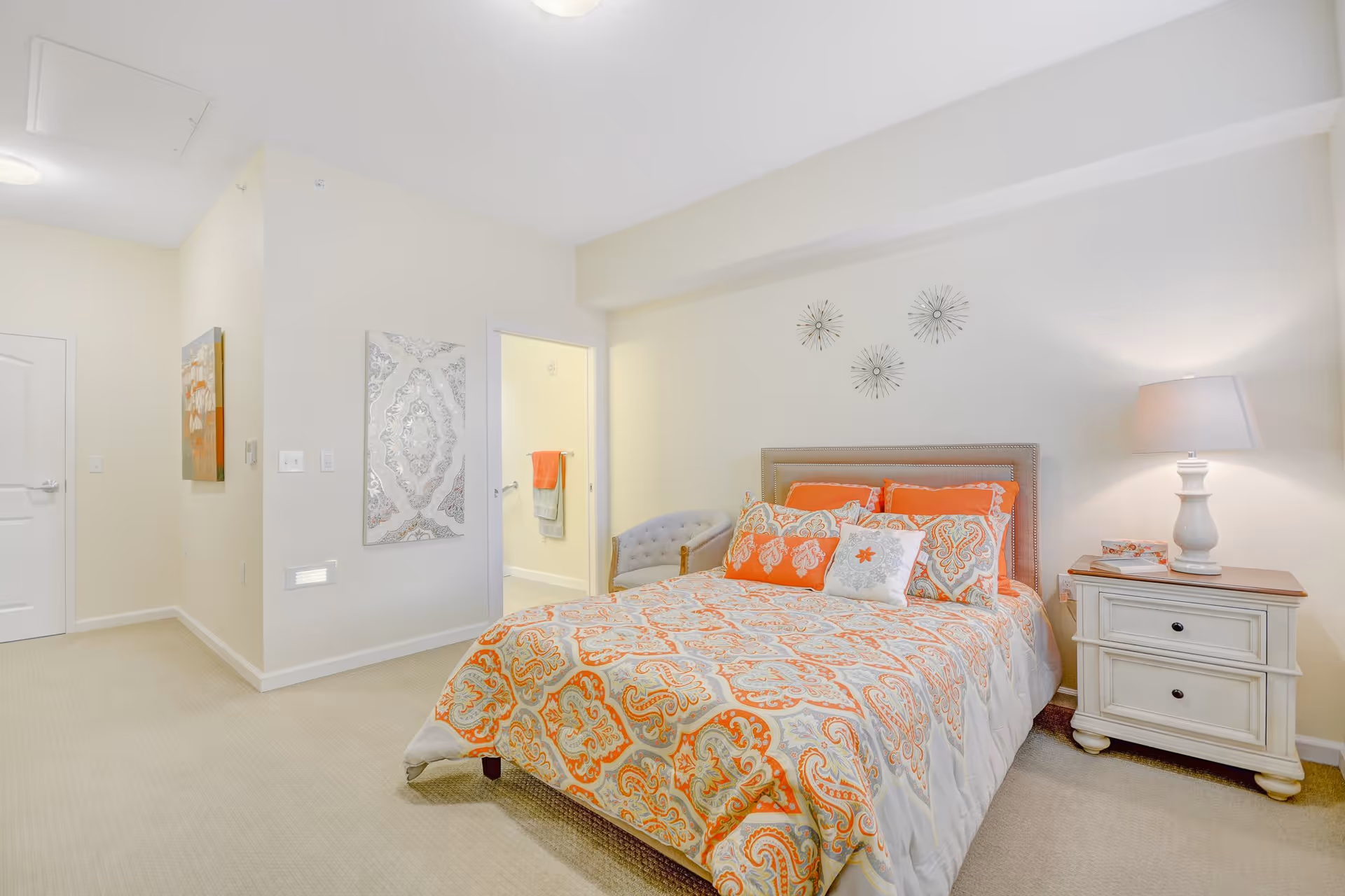 A bedroom with a bed covered in an orange and gray patterned comforter and multiple matching pillows. There is a white nightstand with a lamp and some books on the right side of the bed. A gray upholstered chair is placed near the open doorway leading to a bathroom with towels hanging on the wall. The walls are light-colored with decorative wall art above the bed and on the adjacent wall.