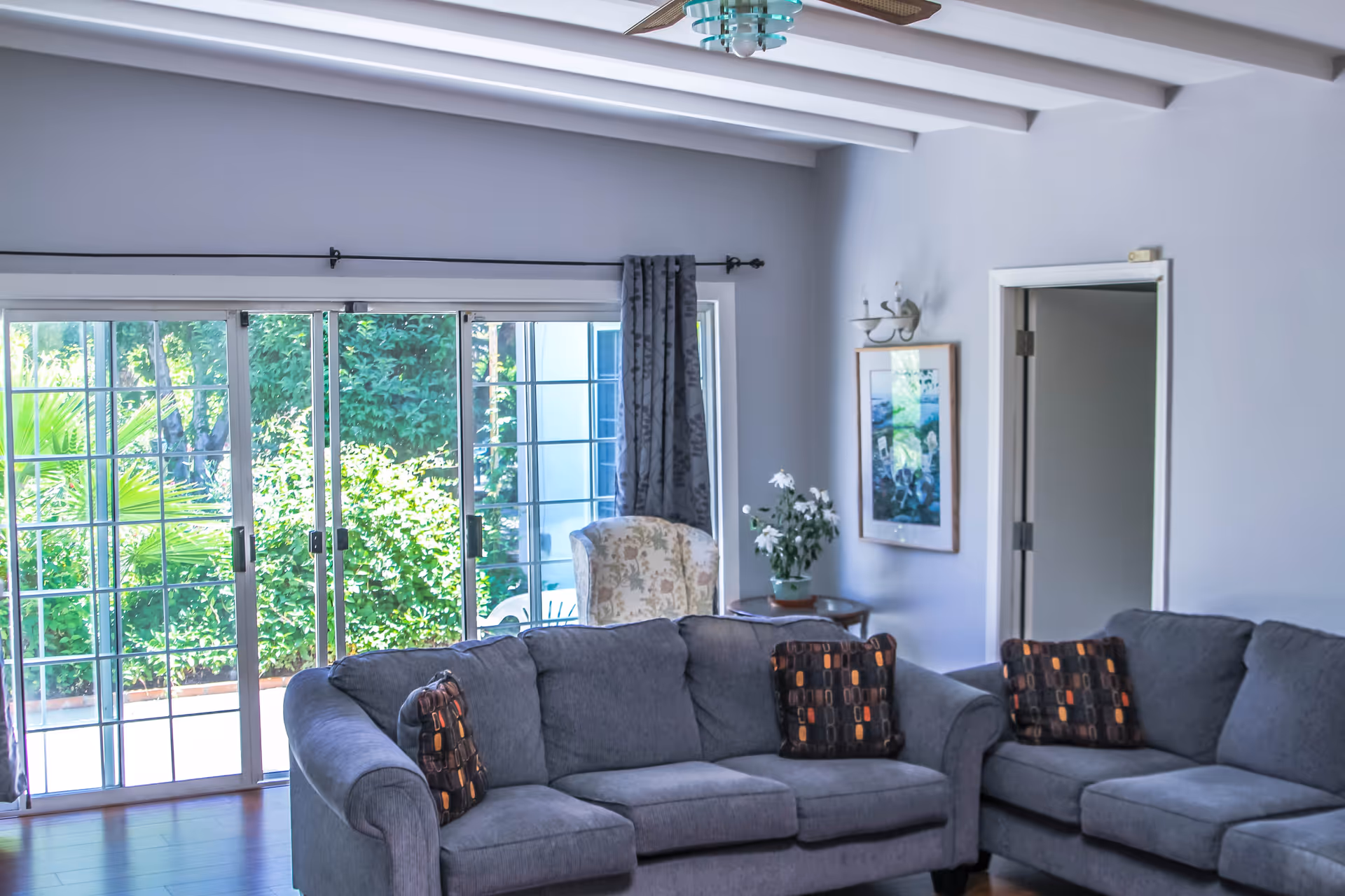 A bright living room with two gray sofas adorned with patterned cushions, a floral armchair near a sliding glass door that opens to a lush green outdoor area, a small round table with a potted plant, and a framed picture on the wall.