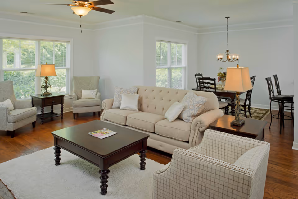 Bright furnished living room with a beige sofa, armchairs, a dark wood coffee table, lamps, and a dining area visible in the background.