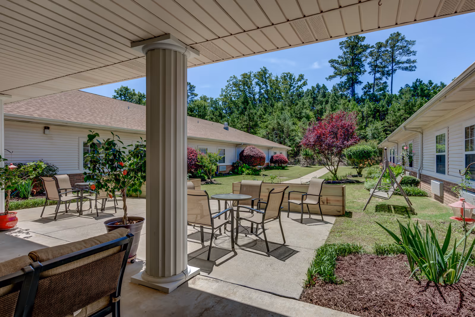 Outdoor patio area at Garrett Woods Senior Living Community with several chairs and tables arranged on a concrete surface. The patio is surrounded by well-maintained landscaping including potted plants, bushes, and trees. Two single-story buildings with white siding and brick accents frame the patio, and a swing is visible on the grass. The sky is clear and blue.