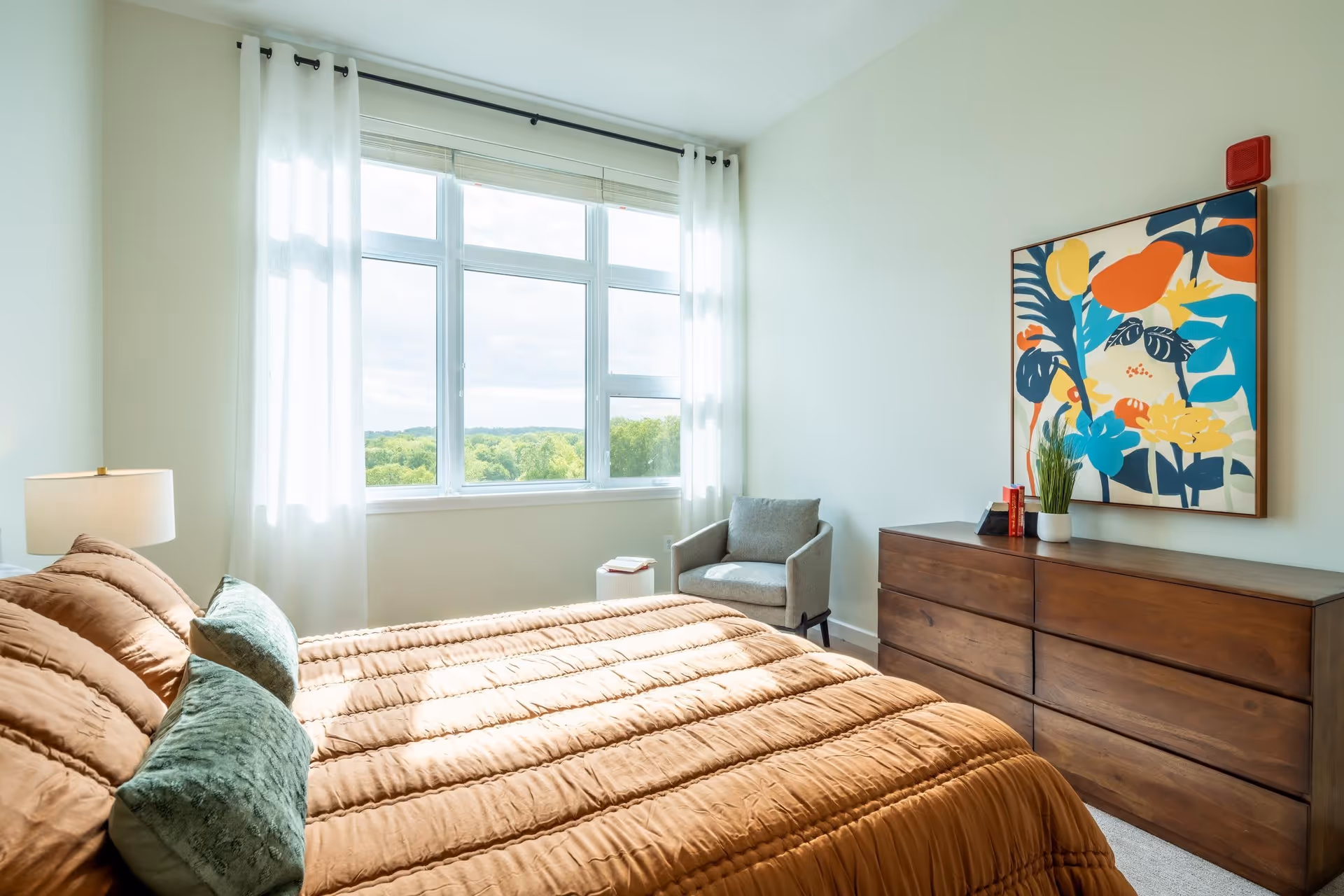 A bright bedroom with a large window covered by sheer white curtains, a bed with a brown comforter and green pillows, a wooden dresser with a colorful abstract painting above it, a gray armchair in the corner, and a lamp on a bedside table.