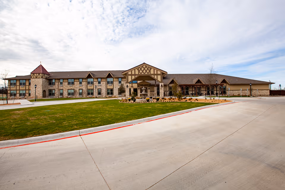 Front exterior view of The Villages of Windcrest, a two-story building with stone and beige siding, a covered entrance with a peaked roof, and a well-maintained lawn and driveway in front.