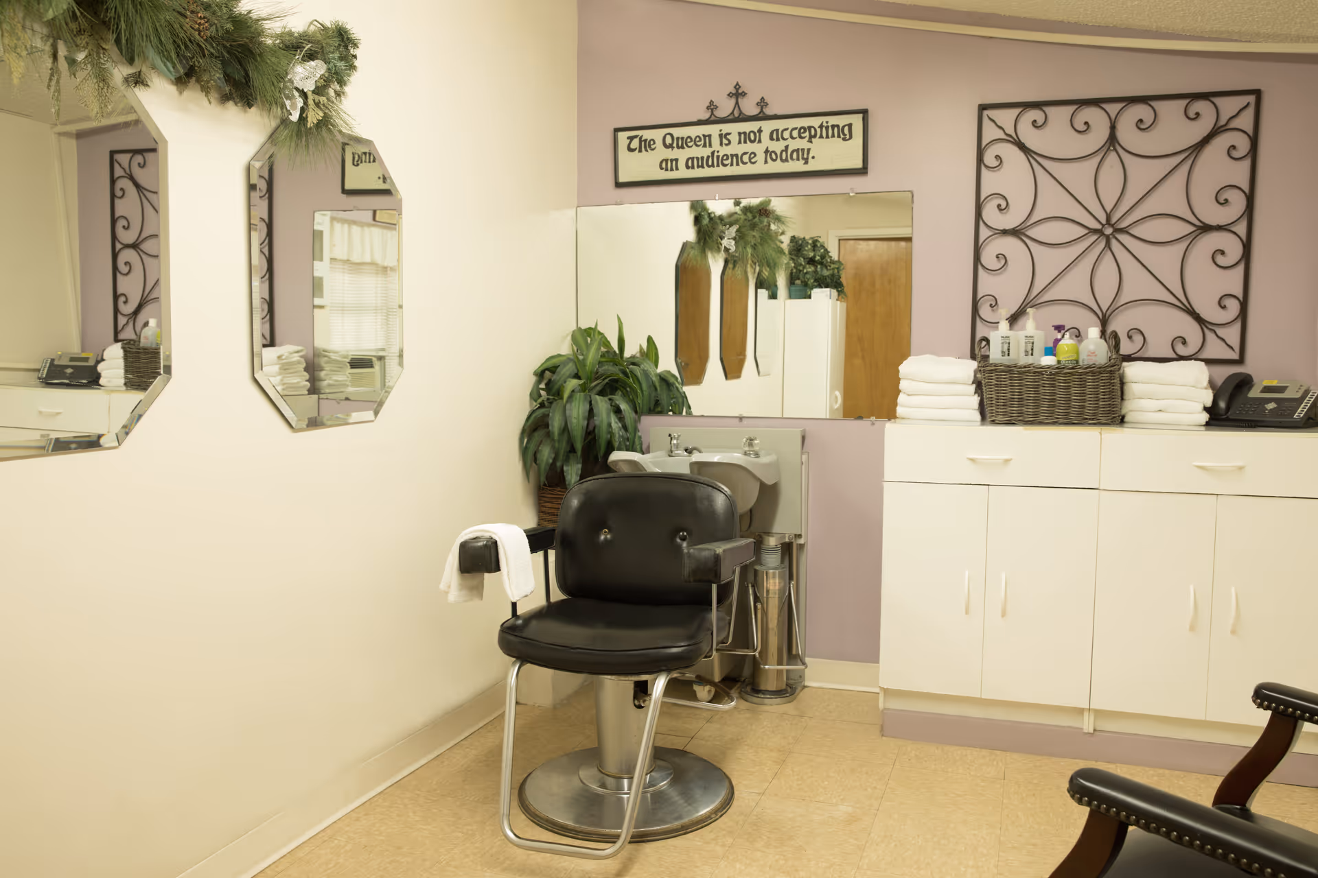 Interior view of a small salon or grooming area with a black salon chair in front of a sink. There are mirrors on the wall, a cabinet with neatly folded white towels, various bottles of hair or skin products in a basket, a telephone, and decorative wall art. A sign above the mirror reads, 'The Queen is not accepting an audience today.'