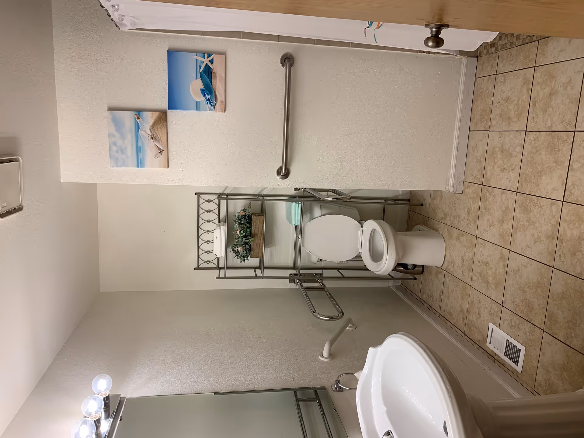 A clean bathroom with a white toilet, a white pedestal sink, and a metal shelving unit holding towels and a plant. The walls are light-colored with two beach-themed pictures hanging above the toilet. The floor is tiled with beige tiles, and there are grab bars installed near the toilet for accessibility.