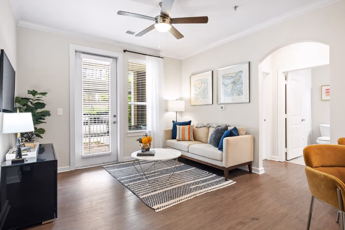 Bright living room with a sofa, throw pillows, round coffee table on a patterned rug, TV console, and glass doors to the outside.